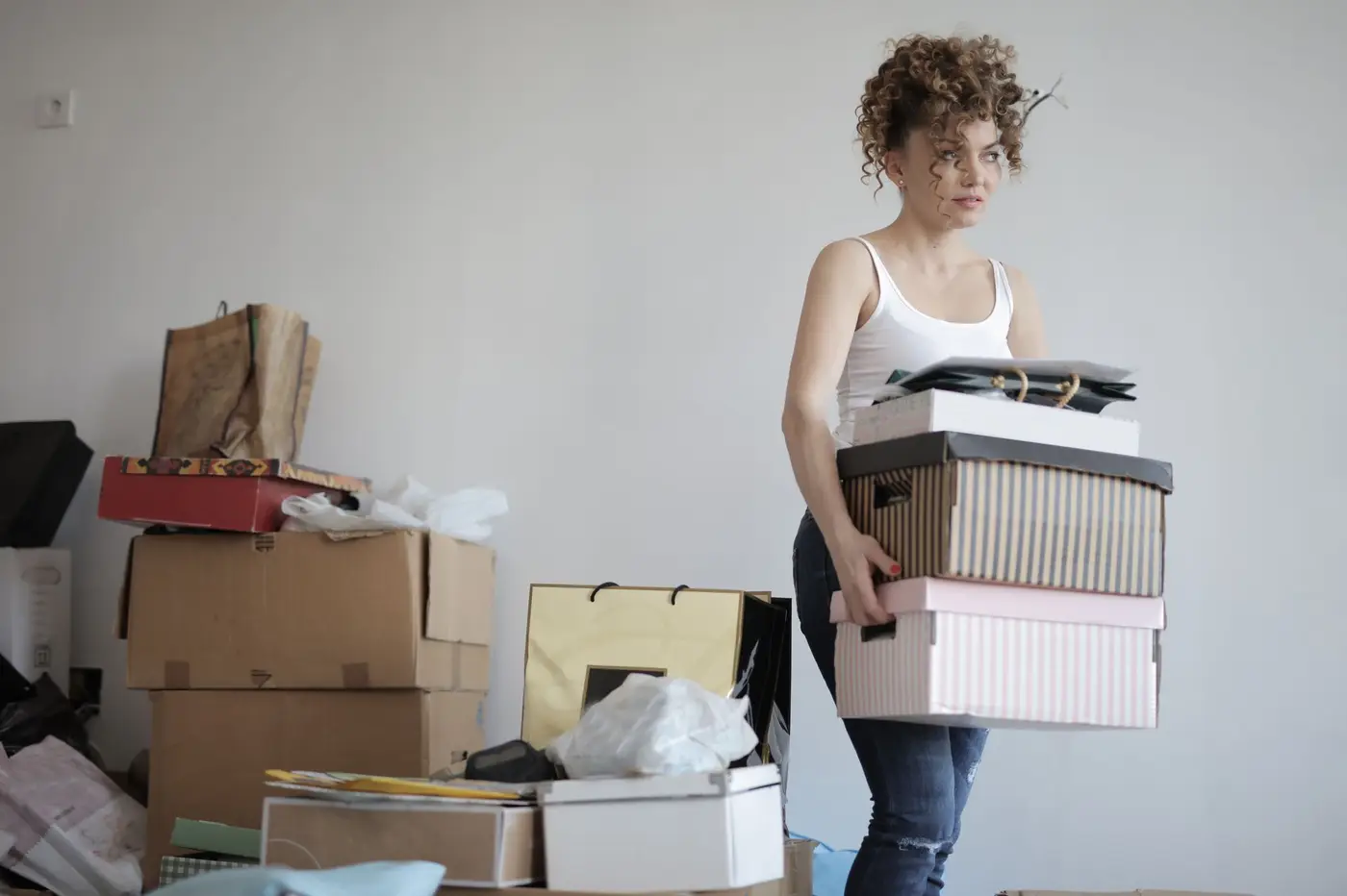 A woman carrying a stack of boxes in a room full of moving boxes.